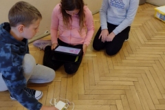Dwie dziewczynki i chłopczyk wpatrują się w ekran tabletu, który trzyma na kolanach jedna dziewczynka. przed nimi na podłodze robot.