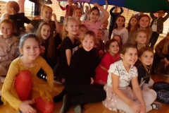 Grupa uśmiechniętych dziewczynek siedzi na sali gimnastycznej pod kolorową chustą.