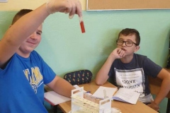 Dwaj chłopcy siedzą w sali przy stoliku, jeden pokazuje próbówkę z czerwoną cieczą
