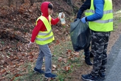 Chłopiec w różowej bluzie i kamizelce odblaskowej wrzuca śmieci do worka trzymanego przez dwóch chłopców w kamizelkach odblaskowych.