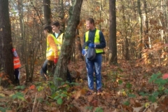 Troje uczniów w kamizelkach odblaskowych spaceruje po lesie.