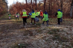 Grupa dzieci w odblaskowych kamizelkach wśród wysokich drzew liściastych szuka śmieci.