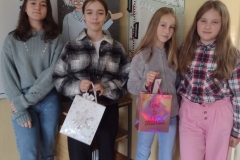cztery dziewczyny w kolorowych ubraniach stoją na tle postaci Pitagorasa i przyrządów matematycznych. W rękach trzymają kolorowe torebki.