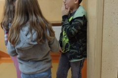 Dwie dziewczyny z długimi włosami w szarych bluzach rozmawiają z chłopcem w bluzie moro  i okularach.
