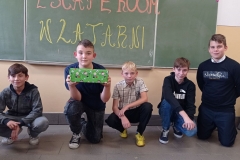 Pięciu chłopców klęczy na tle zielonej tablicy. Jeden z nich trzyma w rękach zielone pudełko.