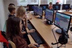 Dzieci w kolorowych ubraniach siedzą w pracowni komputerowej i pracują . Na monitorach platforma code.org