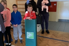 Chłopiec w czerwonym dresie stoi oparty o duże, zielone pudło. Za nim kobieta i mężczyzna z małym dzieckiem na rękach