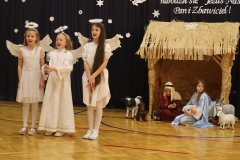 Trzy dziewczynki przebrane za anioły śpiewają kolędę. Za nimi szopka, a w niej Maryja i Józef