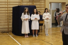 Trzy dziewczynki ubrane na biało stoją trzymając w rękach świeczki. Każda nich ma na piersiach napis wiara, nadzieja lub miłość