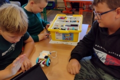 Dwóch chłopców układa program na czarnym tablecie stojącym na stoliku.  Na stoliku znajduje się robot oraz żółte pudełko. W tle inni uczniowie. .
