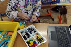 Dziewczynka w kolorowej bluzie składa robota z klocków lego. Przed nią na stoliku  leży żółte pudełko oraz małe białe pudełka z przegródkami w których są klocki.