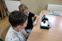 Dwóch chłopców siedzi w ławce, przed nimi stoi mikroskop