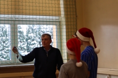 Dwaj chłopcy w mikołajkowych czapkach stoją przy nauczycielu, który pokazuje publiczności efekt doświadczenia