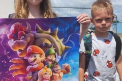 Dziewczynka stoi na polu i trzyma plakat z filmu Super Mario Bros, obok stoi chłopiec, w tle widać chmury.