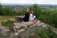 Na ruinach zamku siedzą dwie dziewczynki przytulone plecami i uśmiechają się do fotografującego. W tle widać panoramę miasta.
