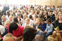 Uczniowie i nauczyciele siedzą na sali gimnastycznej podczas uroczystej akademii