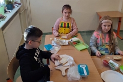Dzieci siedzą przy stolikach i przygotowują produkty do zrobienia kanapek.