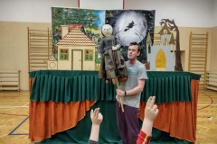 Aktor stoi na tle dekoracji, w rękach trzyma kukiełkę
