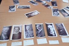 na stoliku uczniowskim, w jednym rzędzie,  leżą wizerunki sześciu mężczyzn, przy każdym zdjęciu biała karteczka z imieniem i nazwiskiem. Nieco dalej kilkanaście innych zdjęć, które leżą w nieładzie.