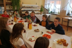 Chłopcy i dziewczynki siedzą przy stole przykrytym białym obrusem. Na stole widać mandarynki, napoje i ciasta