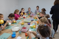Dzieci siedzące dookoła stołu zdobią farbami i brokatem bombki.