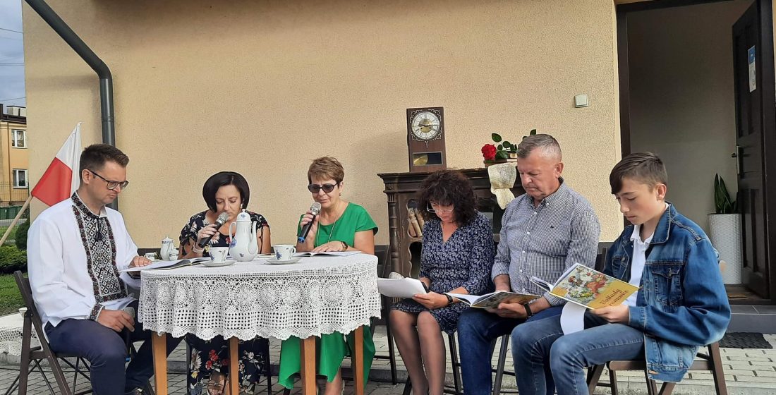 Sześć osób siedzi wokół stolika nakrytego białą serwetą, dwie kobiety trzymają w ręku mikrofony