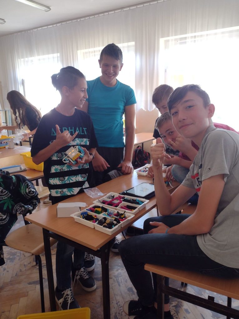 Pięciu chłopców siedzi przy stoliku i buduje z klocków lego postać skoczka. Chłopcy patrzą w obiektyw aparatu. Przed nimi na stoliku rozłożone, posegregowane klocki lego w białych pojemnikach oraz czarny tablet.  