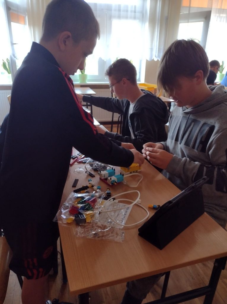 Trzech chłopców buduje robota według instrukcji. Przed nimi na stoliku rozłożone elementy zestawu lego prime oraz czarny tablet. 