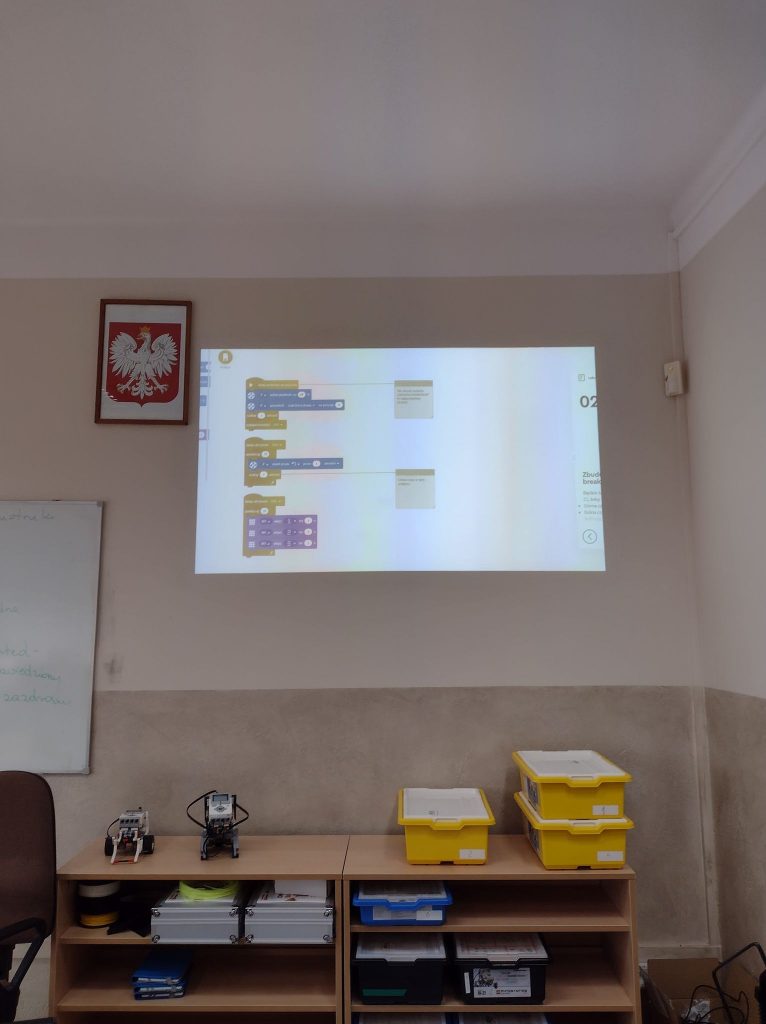 Sala pracowni komputerowej. Na ekranie projektora wyświetlony program. Pod ekranem stoi półka a na niej żółte, niebieskie i czarne pudełka pudełka z klockami lego. dwa zbudowane roboty z ego EV3. 