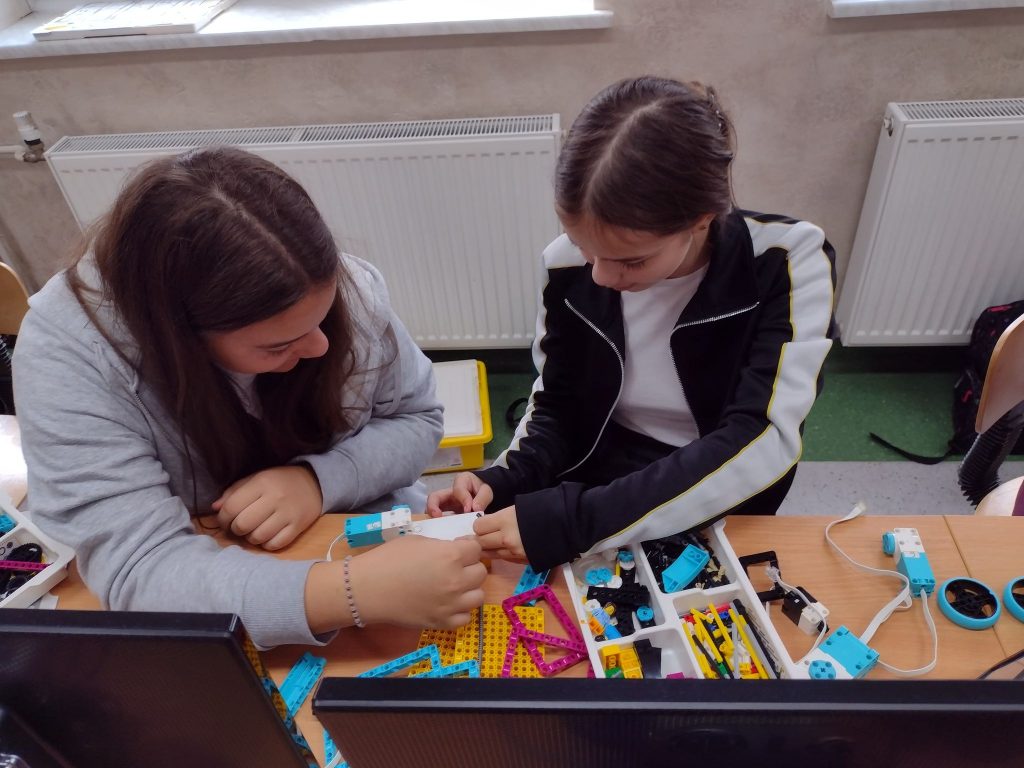 Dwie dziewczyny budują model z klocków lego prime. Przed nimi na stoliku klocki w białym pudełku.