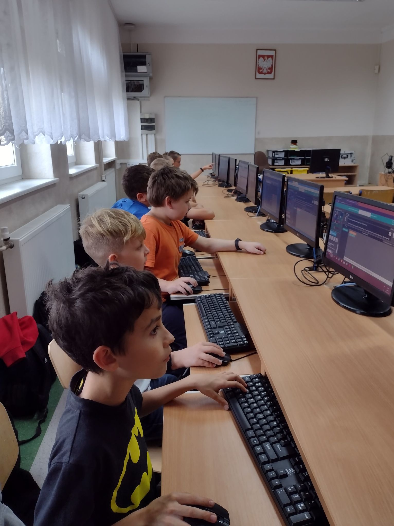 Dzieci w kolorowych ubraniach pracują przy komputerach. Na ekranach komputerów platforma code.org